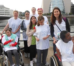 5 staff and 4 service users from the Anne Sullivan centre pose in front of the River Liffey in Dublin docklands after completing the Dublin docklands run. They are wearing matching white t-shirts. 2 of the service users are using a wheelchair.