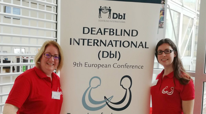 Deirdre on left and Heather on right are wearing red t-shirts and standing beside a large deafblind International sign in white colour and deafblind international logo.