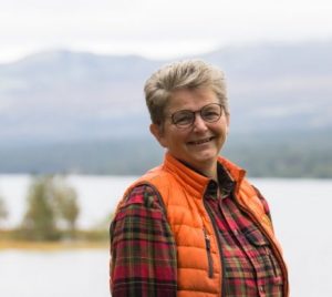 Image of Grete Steigen, standing outside in an orange and red coloured clothing. There are mountains and body of water in the background.
