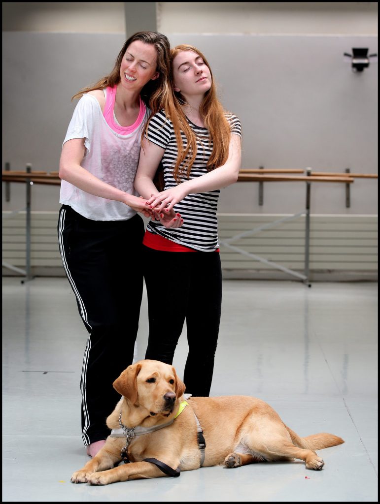 Image of 2 dancers joining hands side by side with a golden retriever dog at their feet.