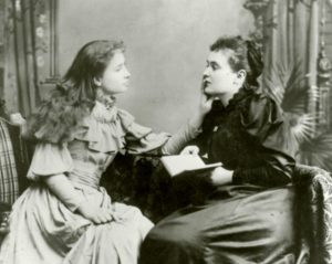 Anne Sullivan on right is reading aloud to Helen Keller. Keller's left hand touches Sullivan's lips to feel the vibrations of Sullivan's spoken words using the tadoma method.