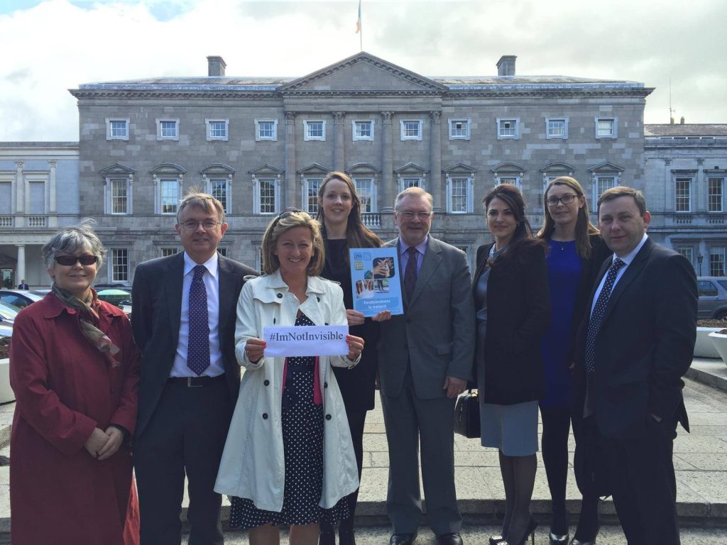 A group of 8 people are standing in front of  government buildings holding up a sign saying "#ImNotInvisible" and a report on deafblindness.